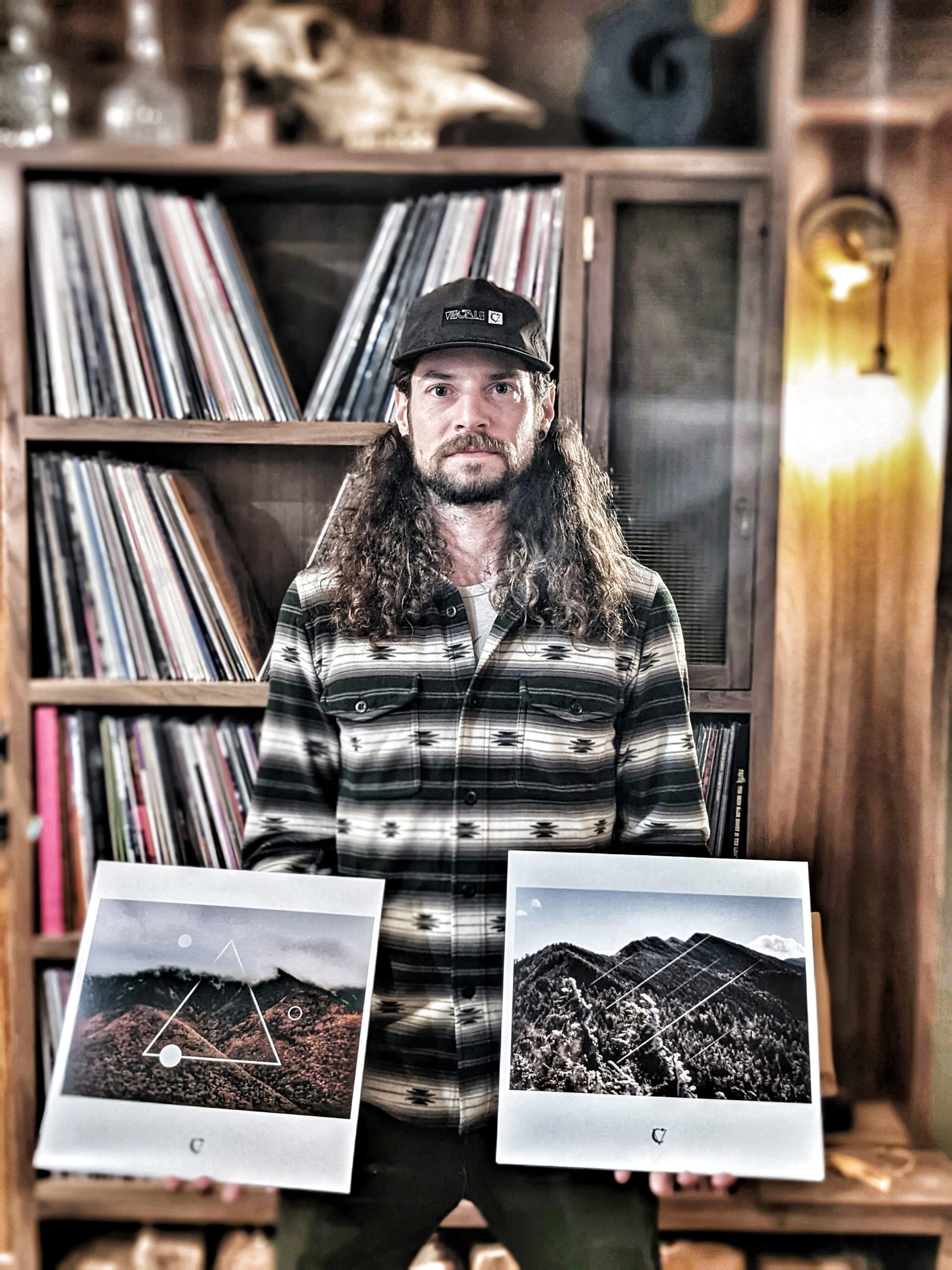 Tim Gormley, wearing a black cap and a button-down shirt, holds two album covers while standing in front of his record collection.