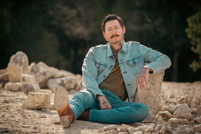 Zach Meadows, wearing jeans and a denim jacket, sitting on a rock outcropping.